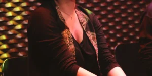 Seated vocalist with curly hair performing into a microphone, eyes closed, in warm red lighting against a textured patterned wall.