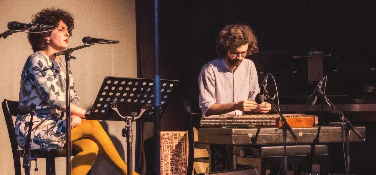 Seated vocalist and santur player performing on stage at Sargfabrik during the Nowruz festival (Vienna, March 2018); singer at left with floral dress and yellow tights, santur player tuning at right.
