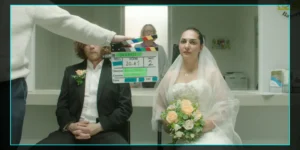 Behind-the-scenes monitor frame from Shahid: a clapperboard marked “Scene 20.A1, Take 2” is held in front of a bride in a white gown holding peach roses, seated beside a man in a suit with a boutonnière inside a registry-office setting; a crew member stands behind glass, with on-screen camera data (FPS 25, 5600K) around the border.