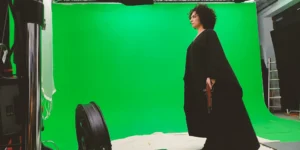 Behind the scenes of Shahid: a performer in a black robe stands side-on on a treadmill before a green screen, holding a prop gun; studio lights, a floor fan, cables, and a ladder are visible around the set.
