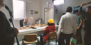 Behind the scenes of Shahid: crew members with boom poles and a clapperboard surround a set dressed as a government service counter; a performer in a yellow beanie and red coat sits facing a clerk behind glass under bright LED panels.