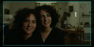 On-set monitor frame from Shahid: two curly-haired women lean together and smile softly toward the lens in a dim apartment kitchen/living room; a crew member stands by a table in the background; camera HUD shows “FPS 25,” “5600K,” and “STBY.”