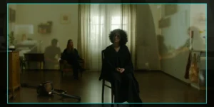 On-set monitor frame from Shahid: a curly-haired woman in a black robe sits on a chair in a dim apartment, another figure seated behind near sheer curtains; a vacuum lies on the floor and soft projections blur across the walls, with camera HUD showing “REC.”
