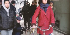 Behind the scenes of Shahid: a woman in a yellow beanie and red coat stands with a vintage bicycle in a cobblestone passage while crew members and a camera rig set up behind her.