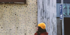 Film still from Shahid: a woman in a yellow beanie and red coat pauses with a vintage bicycle beside a tiled wall and a parking sign on a city street corner.