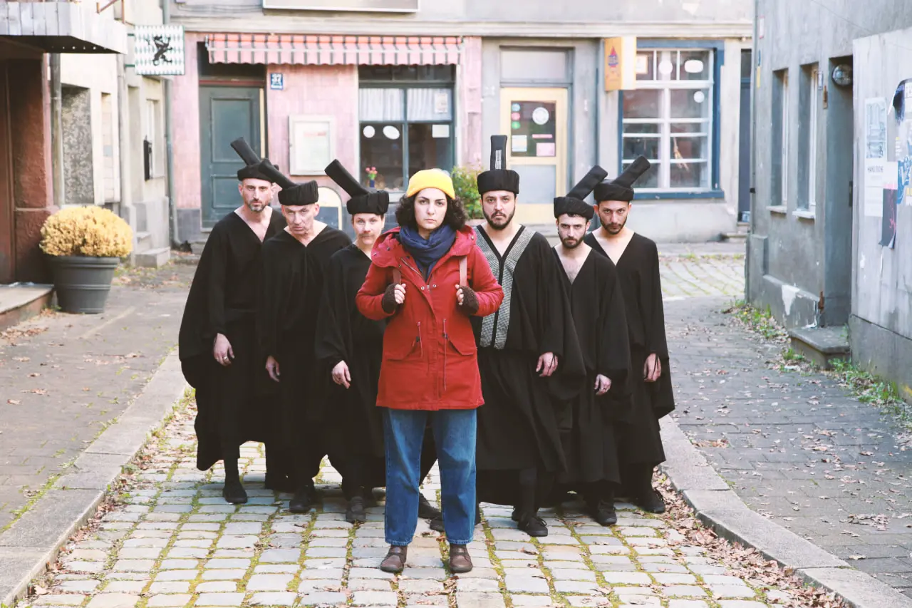 Film still from Shahid: a woman in a yellow beanie and red coat walks ahead of a solemn black-robed chorus with tall headpieces on a cobblestone street lined with storefronts.
