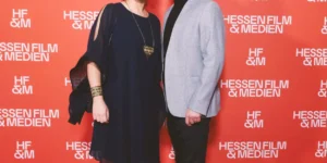 Red carpet photo for Shahid: two guests pose in front of a red “Hessen Film & Medien” step-and-repeat—one in a navy dress with statement jewelry, the other in a light grey blazer and black turtleneck.
