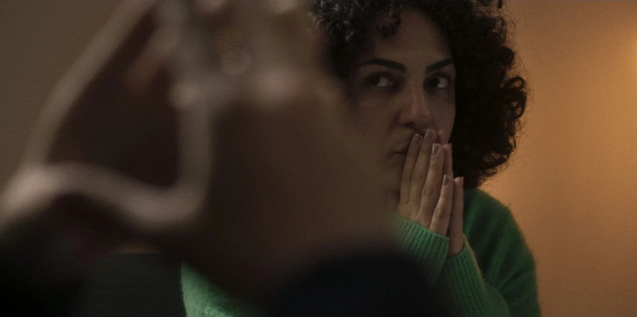Film still from Shahid: close-up of a curly-haired woman in a green sweater, hands pressed to her lips, glancing sideways in a dim, warm room with a blurred foreground.