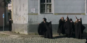 Film still from Shahid: on a sunlit cobblestone corner, a woman in a black robe lifts her arms before a worn gray wall, while a chorus of robed performers raises hands at right; a passerby stands near a parking sign.