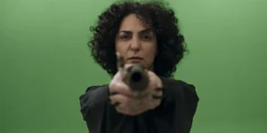 Film still from Shahid: a curly-haired woman in a black robe aims a rifle straight at the camera against a green-screen backdrop.