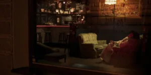 Film still from Shahid: view through a window into a dim bar; a woman in a light dress reclines on a floral armchair with a drink, vintage couches and a chalkboard bar glowing warmly.