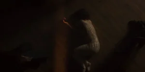 Film still from Shahid: overhead view of a woman curled on a dark wooden floor, her body covered with handwritten map-like markings; a thin strip of warm light cuts across, with dark garments at the edges.