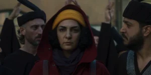 Film still from Shahid: close-up of a woman in a yellow beanie under a red hood, staring ahead as a black-robed chorus surrounds her; blurred hands lifted in the background.