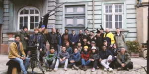 Group photo of the Shahid cast and crew on location: dozens pose in front of a teal building on a cobblestone street, with a camera rig at left.
