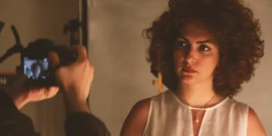 a photographer frames a curly-haired woman in a white blouse; her face appears on the camera screen in a studio setup