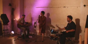 Live music setup for Arash // Heimkehrer: a vocalist stands at a microphone with a tambourine, flanked by a seated electric guitarist (left) and a drummer at a small kit (right), in a warm, candlelit room with a purple wash on a cracked plaster wall.