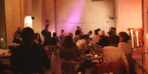 Wide view of a candlelit café-style room during Arash // Heimkehrer: seated audience gathers at small tables while a vocalist, guitarist, and drummer perform under a purple wash on the back wall.