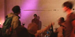 Live music at Arash // Heimkehrer: a vocalist stands at a microphone against a purple-lit plaster wall while audience members blur past in the foreground; a drummer and instruments are partially visible at right.