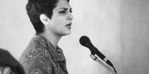 Black-and-white close-up of a short-haired vocalist in a patterned dress singing at a side microphone, softly lit with shallow depth of field.