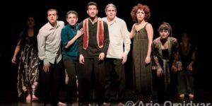 Stage photo from Nodbeh: seven performers in mixed costumes stand shoulder to shoulder under a hard spotlight, staring forward on a black stage.