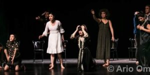 Stage photo from Nodbeh: a line of performers spreads across the stage—some kneeling, some standing barefoot—reaching and crying out with expressive gestures; scattered chairs and a black background frame the scene.