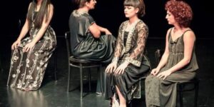 Stage photo from Nodbeh: four performers sit on simple black chairs in a diagonal, wearing dark patterned dresses—one barefoot—gazing in different directions against a black stage.