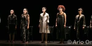 Stage photo from Nodbeh: eight performers stand in a straight line under a tight spotlight, a few barefoot, with empty black chairs behind them against a dark stage.