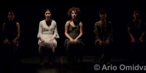 Stage photo from Nodbeh: five performers sit in a row under narrow spotlights—center figures in a white dress and a long green dress—surrounded by deep blackness.