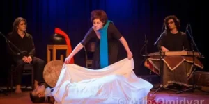 Funeral-ritual tableau in “Siavoshan”: the protagonist lifts a white shroud over a prone figure while two musicians accompany from upstage.