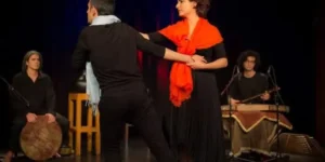 Dance sequence from “Siavoshan,” the protagonist wrapped in an orange shawl partnering a male dancer, with live percussion and santur behind.