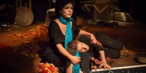 Mourning tableau from “Siavoshan”: the protagonist in a turquoise scarf cradles a fallen body beside a carpet of petals, instruments and musicians dimly lit in the background.
