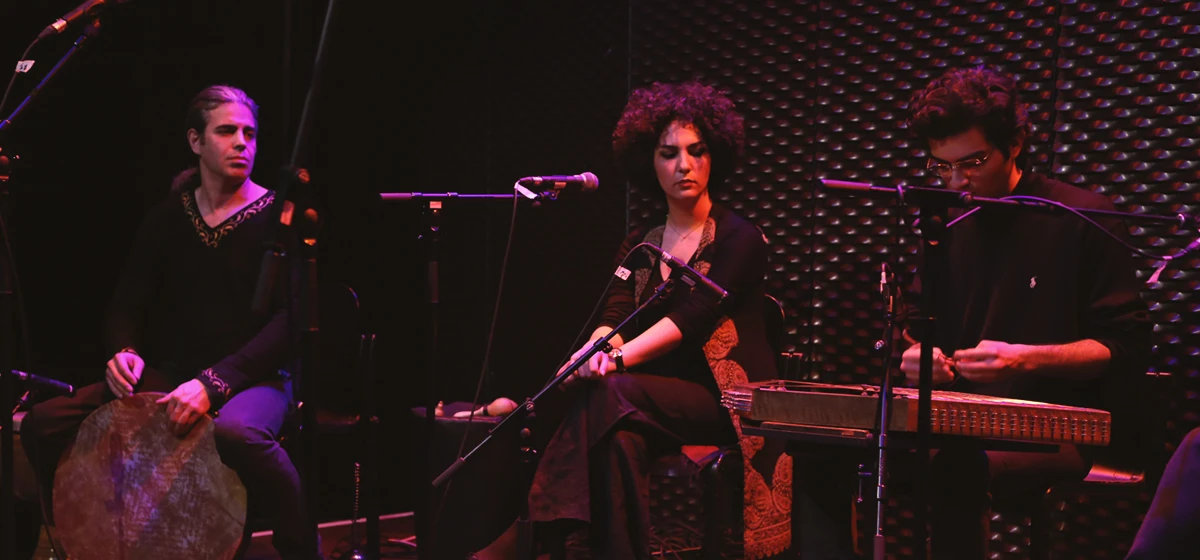 International Women’s Day concert at WUK: trio on stage—a frame-drum player (left), a seated vocalist at the center mic, and a santur player (right)—under magenta lighting against a textured black wall.
