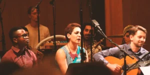 Seated vocalist, Bahark Abdolifard singing into a microphone beside an acoustic guitarist; ensemble players and multiple mic stands in the background at RadioKulturhaus.