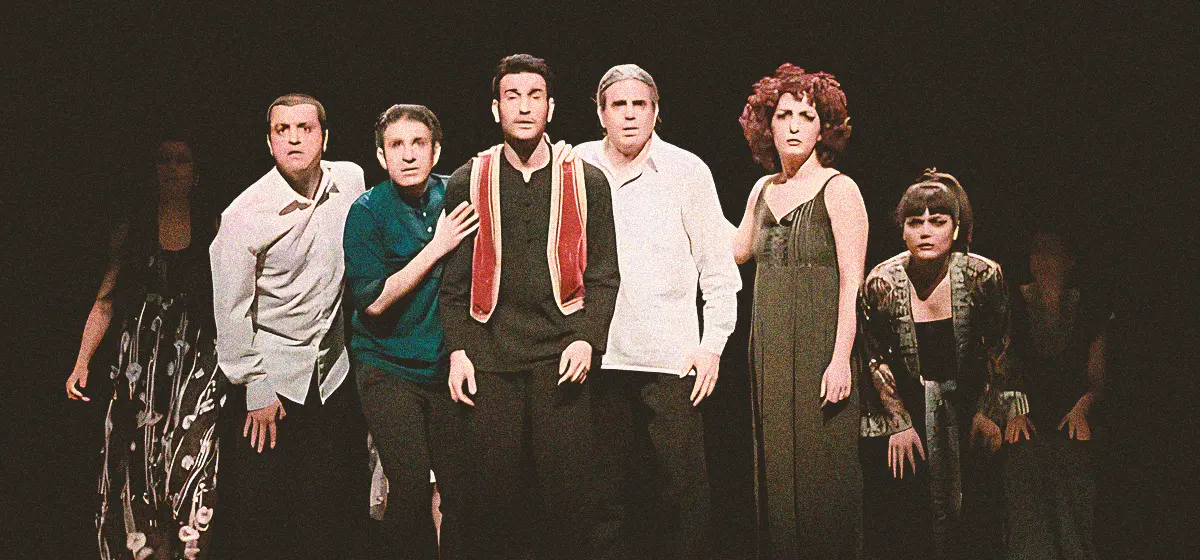 Stage photo: seven performers stand in a tight line under a spotlight, faces intent and urgent, with mixed contemporary costumes against a black background—capturing a dramatic moment in a theatre piece.