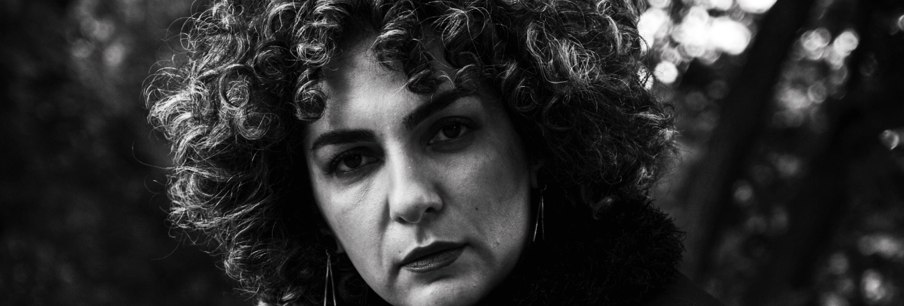 Black-and-white close-up of a woman with curly hair and a serious gaze, trees softly blurred in the background.