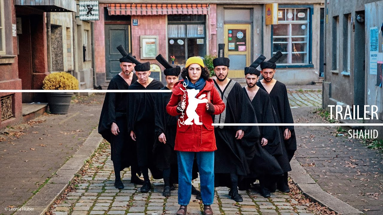 Trailer cover for Shahid: a woman in a yellow beanie and red coat stands on a cobblestone street facing the camera as a black-robed chorus lines up behind her; Berlinale bear logo overlays the center with “TRAILER — SHAHID” text at right.