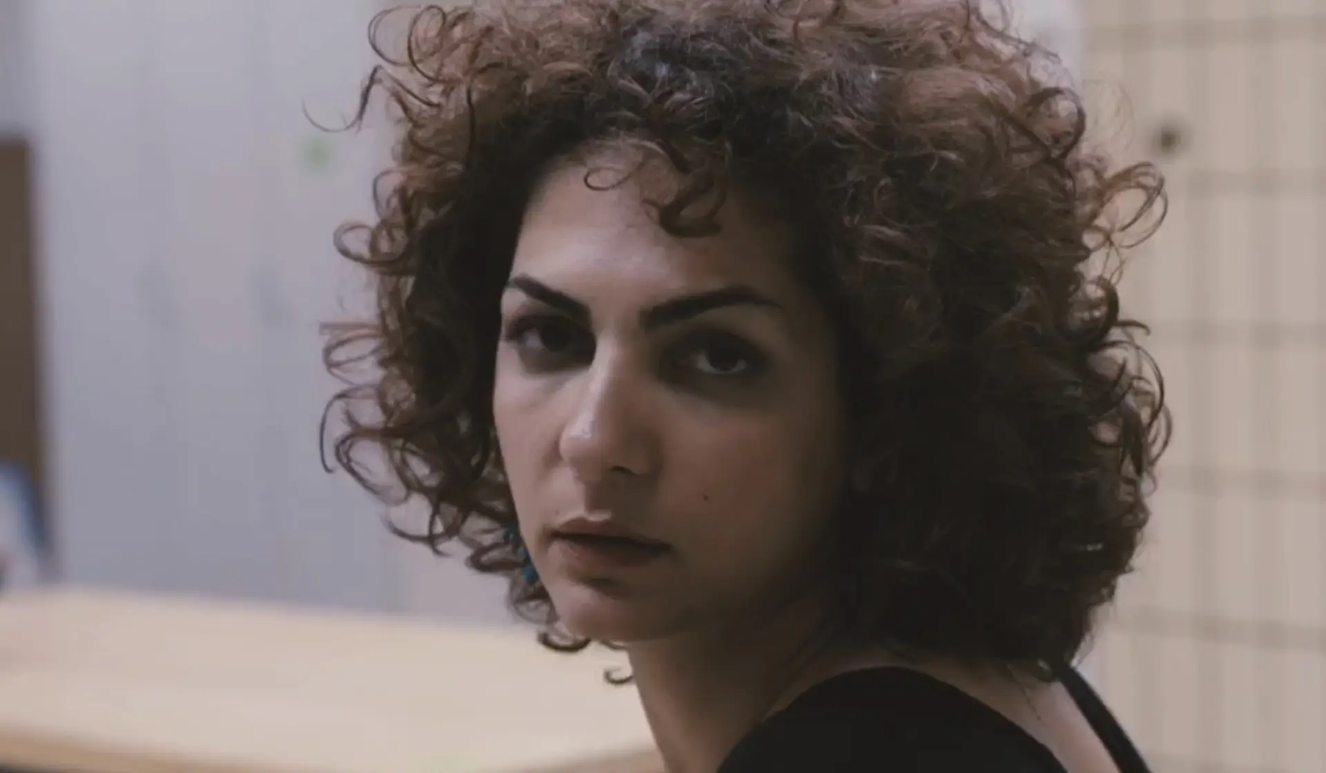 Trailer still from The Photographer, His Lover, His Sister and Her Lover (2017): close-up of a curly-haired woman turning toward the camera, soft light and tiled wall in the background, pensive expression.